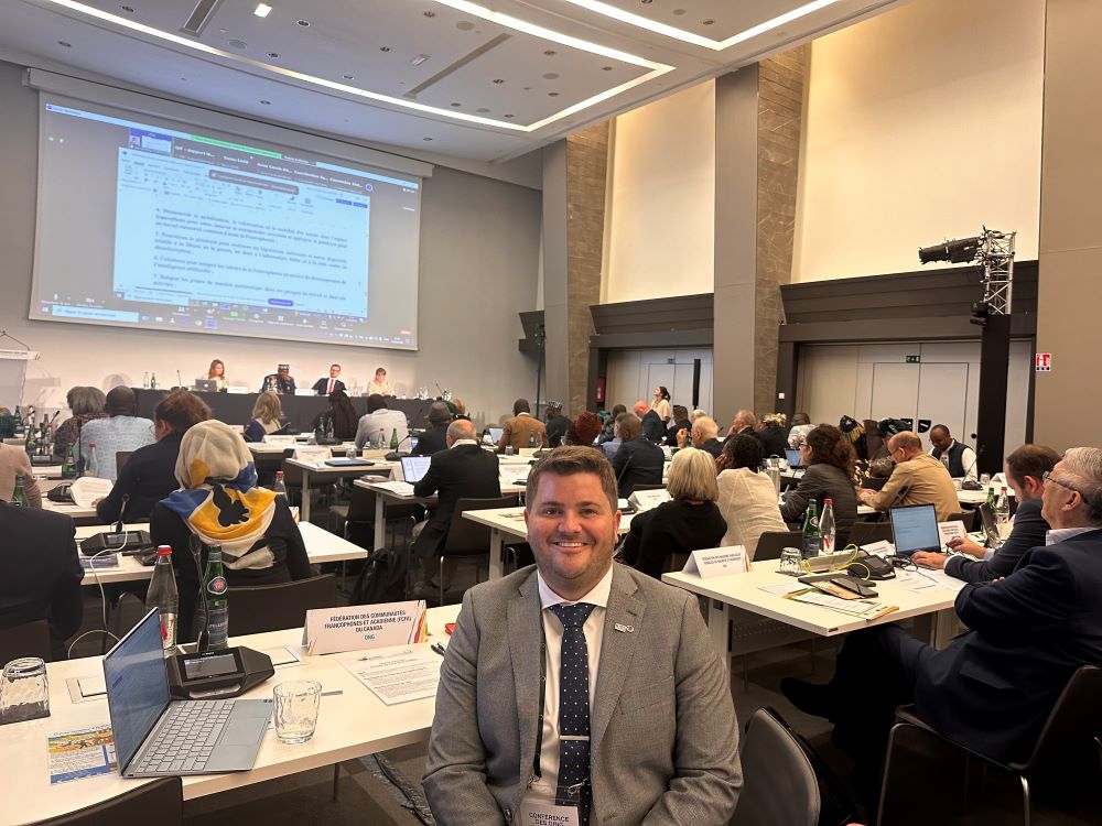 Photo d'un homme en complet assis dans une salle de rencontre en compagnie d'autres personnes faisant face à un écran avec une projection Powerpoint. 